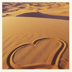 a heart drawn in the sand on top of a beach with dunes and clouds behind it