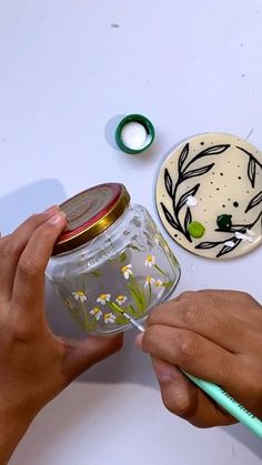 a person is painting a jar with flowers and leaves on the lid, while another hand holds a green toothbrush