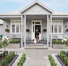 two people sitting on the front porch of a gray house with white flowers and shrubs