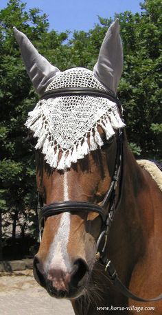 a brown horse wearing a white crochet head cover and bridle with trees in the background