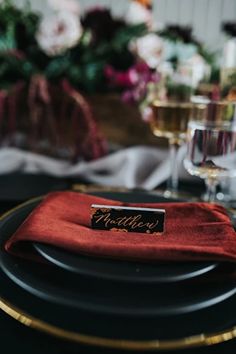 a place setting with red napkins and gold rimmed plates, wine glasses and flowers in the background