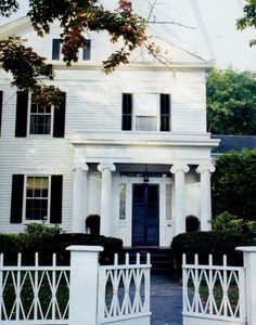 a large white house with black shutters on the front and side doors, surrounded by trees