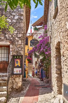 an alley way with stone buildings and purple flowers