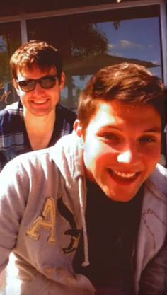 two young men sitting next to each other in front of a glass door and smiling at the camera