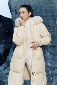 a woman standing in front of a stone wall wearing a white coat and fur collar