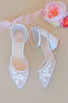 two pairs of white wedding shoes next to pink flowers on a beige surface with ribbon