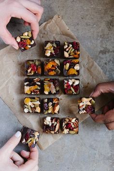 two hands reaching for pieces of food on top of a piece of parchment paper with nuts and cranberries