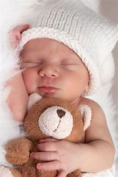 a baby sleeping with a teddy bear in it's arms and wearing a white hat