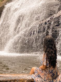 a woman sitting in front of a waterfall with her back to the camera and looking at it