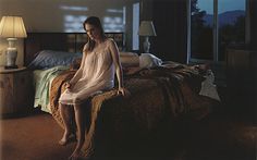 a woman sitting on top of a bed next to a lamp in a room with large windows