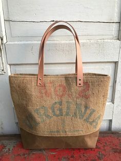 A vintage Virginia burlap peanut sack has been repurposed into this unique sturdy tote bag. It features a dark tan waxed canvas bottom and natural vegetable tanned leather straps.  The interior lining is a medium blue cotton ticking with a 3 sectioned pocket for smaller items. The top is secured with a magnetic snap. It measures a generous 18 in. wide, 15 in. high, and 5 in. deep.  Strap drop measures 10 in. Easily holds all your essentials for the day and is also perfect for travel. Waxed canva Rustic Brown Bags With Leather Handles, Rustic Brown Bag With Waxed Finish, Rustic Bags With Leather Handles For Everyday, Brown Duck Canvas Tote Bag, Brown Canvas Bucket Bag With Leather Handles, Rustic Everyday Tote Bag, Rustic Brown Bag For Everyday Use, Rustic Tote Bag For Everyday Use, Eco-friendly Upcycled Canvas Bag