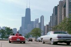 an old car driving down the road in front of a white car and some tall buildings