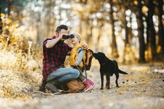 a man and woman are sitting in the woods with a dog looking at something through binoculars