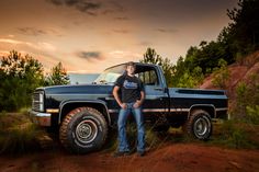 a man standing in front of a truck on a dirt road with trees and bushes behind him