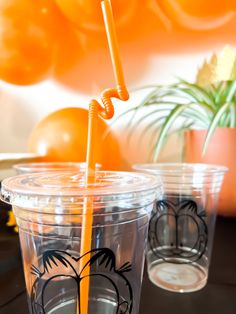 two plastic cups with orange straws in them sitting on a table next to an orange plant