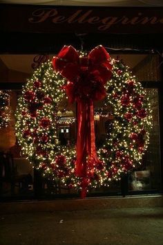 a christmas wreath with red bows and lights in front of a storefront window at night