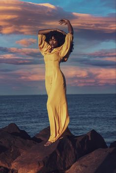 a woman in a long yellow dress standing on rocks near the ocean with her arms outstretched