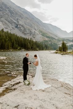 a bride and groom standing on the edge of a lake with mountains in the background