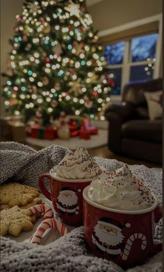 two mugs filled with whipped cream next to cookies and a christmas tree in the background