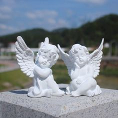 two white angel figurines sitting next to each other on a cement block with trees in the background