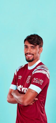 a man with his arms crossed wearing a maroon and white soccer jersey against a blue background