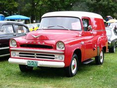 an old red truck parked on top of a lush green field next to other classic cars