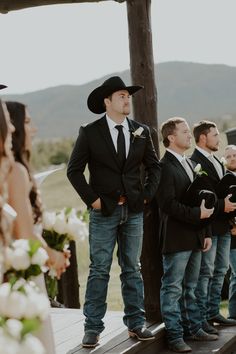 a group of men standing next to each other in front of a wooden structure with white flowers