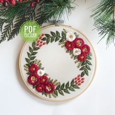 an embroidered wreath with red flowers and greenery is displayed on a white surface next to pine branches