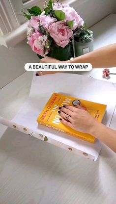 a woman is holding a book in front of a flower pot and an empty box on the floor