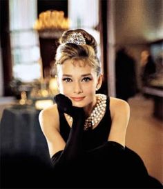 a woman in a black dress with pearls on her head sitting down and looking at the camera