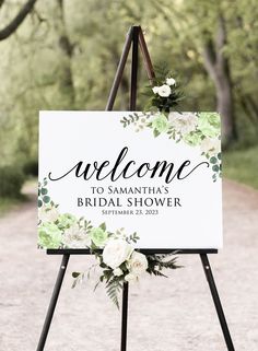 a welcome sign with white flowers on it in the middle of a dirt road surrounded by trees