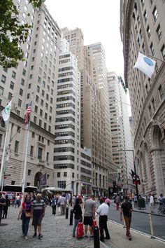 many people are walking down the street in front of tall buildings with flags on them