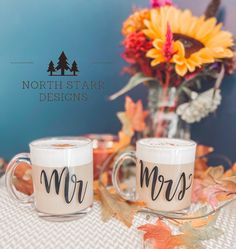 two coffee mugs sitting on top of a table with autumn leaves and flowers in the background