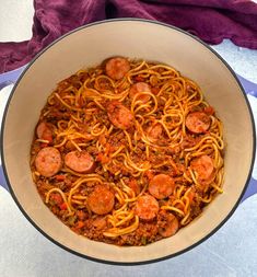 a pan filled with spaghetti and sausages on top of a white table cloth next to a purple towel