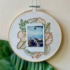 an embroidered photo frame hanging on a wall next to a green leafy plant and a white wall