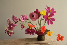 an arrangement of colorful flowers in a vase on a wooden table next to a white wall