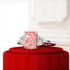 a fancy pink diamond ring sitting on top of a red velvet cushioned stand in front of a white wall