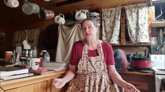 an older woman sitting at a kitchen counter with pots and pans hanging from the ceiling