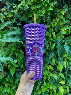 a hand holding up a purple cup in front of some green plants and bushes with a sticker on it