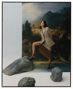 a woman sitting on top of a large rock next to a painting with mountains in the background