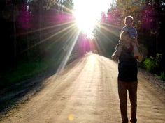 a woman holding a baby walking down a dirt road with the sun shining behind her