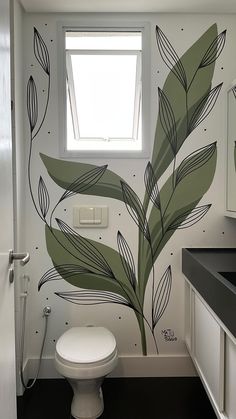 a white toilet sitting under a bathroom window next to a sink and counter top with a plant painted on the wall behind it