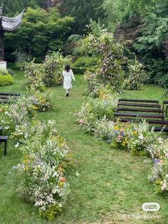 a woman walking through a lush green park filled with lots of trees and flowers next to benches