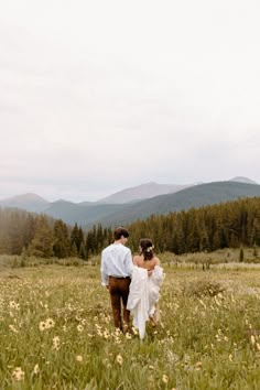 a bride and groom walking through a field with mountains in the background