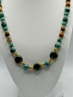 The beads are predominantly in shades of turquoise and brown with a glossy finish, and some have a marbled effect that adds depth and interest to their appearance. Between the larger beads are ornate gold-toned spacer beads with intricate designs, contributing to the necklace's luxurious look. The combination of the earthy brown tones with the vibrant turquoise creates a visually striking contrast. This necklaceswould appeal to those with a taste for bold, statement jewelry that combines natural Elegant Turquoise Agate Beaded Necklaces, Polished Malachite Round Beaded Necklaces, Malachite Gemstone Beaded Necklaces, Round Malachite Beaded Necklace, Beaded Malachite Round Bead Necklaces, Gold Jade Beaded Necklaces With Round Beads, Gold Jade Round Beaded Necklaces, Turquoise Malachite Necklaces With Round Beads, Gold Agate Beaded Necklaces