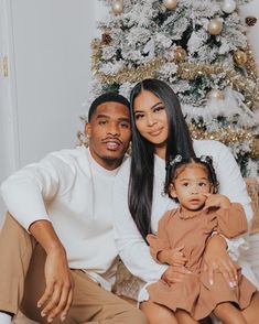 a man and woman sitting next to a christmas tree with a baby in front of them