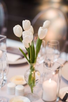 white tulips are in a vase on a table with candles and wine glasses