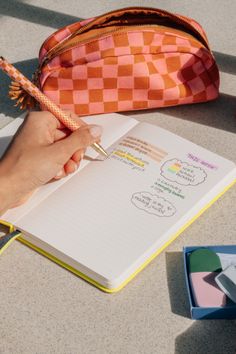 a person holding a pen and writing on a notebook next to an orange checkered bag