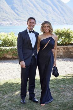 a man and woman in formal wear standing next to each other on the grass with mountains in the background