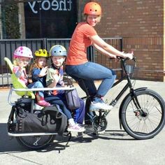 a woman riding on the back of a bike with two small children in front of her